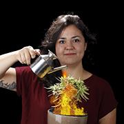 woman standing in red shirt facing forward, Caucasian, holding a watering can over a plant